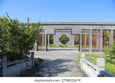 A broad paved path leads through a circular arch in a white wall with red metal window grilles. Trimmed hedges, shrubs, a small bridge, and clear sky create a formal garden entrance with symmetrical lines. The repeated openings, masonry texture, and greenery create a balanced architectural garden composition.