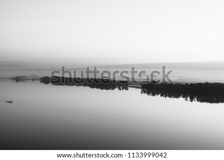Broad river flows along diagonal shore with silhouette of forest and thick fog in grayscale. Tree drifts with flow. Minimalistic monochrome landscape of majestic nature. Morning milky atmosphere.