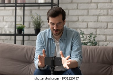 Broadcasting life. Confident male coach tutor sit on sofa in front of phone on holder use gadget as webcam record training webinar. Young man blogger talk speak before cell camera shoot video review