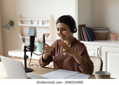 Broadcasting online. Artistic indian woman in headphones sit by desk with laptop speak using professional microphone record audio podcast. Young lady business coach shoot video training at home studio - Powered by Shutterstock