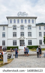 Broc, Switzerland - July 27, 2019: People Waiting In Front Of The Building Of The Famous Cailler Chocolate Factory. Visitors In The Outside Courtyard. Tour And Museum. Swiss Chocolate. Vertical Photo.