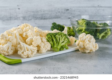 Broccoli florets in a glass bowl, and cauliflower florets close-up on a cutting board on kitchen table - Powered by Shutterstock