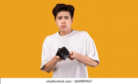 I'm Broke. Portrait of sad asian bankrupt showing his empty wallet and looking at camera. Upset crying male model standing isolated over orange studio background wall. Poverty Concept.
