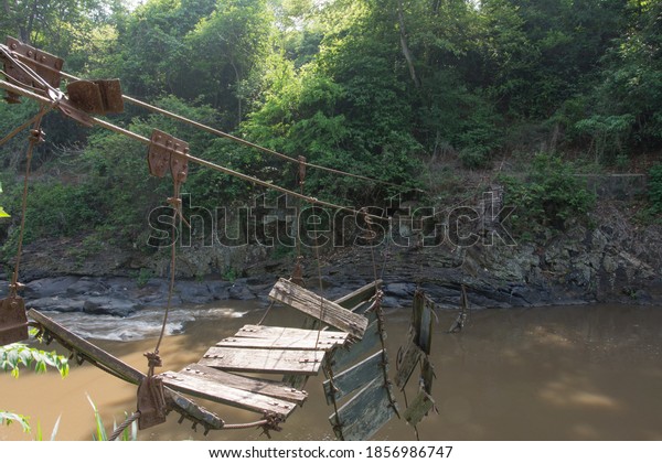 Broken bridge made from wood and steel\
rope over a mountain river in a tropical\
forest.