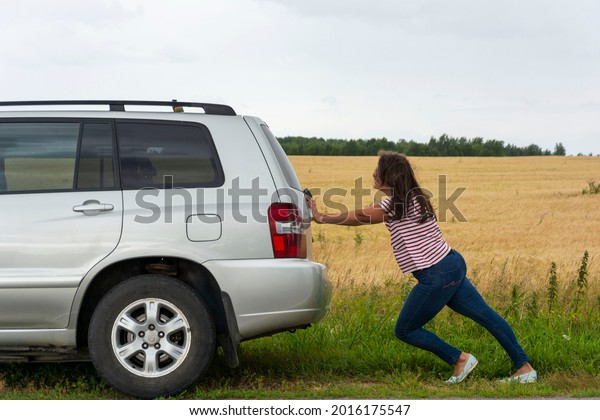 A\
broken car. A young woman pushes a broken car on the road, a\
breakdown, against the background of a yellow\
field.