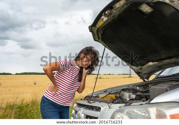 A broken car. A young woman stands on the road by a\
broken car against the background of a yellow field. I need help.\
Helplessly looks under the hood. A phone call to help with a broken\
car
