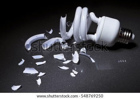 A Broken Compact Fluorescent CFL bulb lays shattered in shards of glass on a hard surface with a dark background