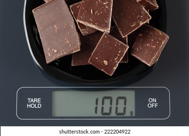 Broken dark chocolate in a black plate on digital scale displaying 100 gram.