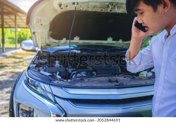 A broken\
down car, engine open and smoking, driver looking at the engine and\
using smartphone call for\
service.