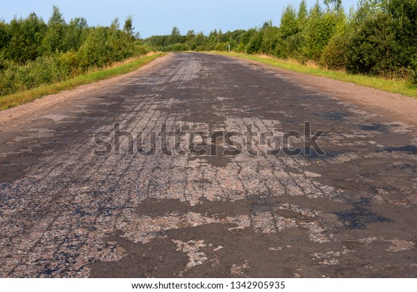 Broken empty asphalt country road surrounded by\
trees in sunny summer day. Road with holes in the green wood. A\
hole in the road.