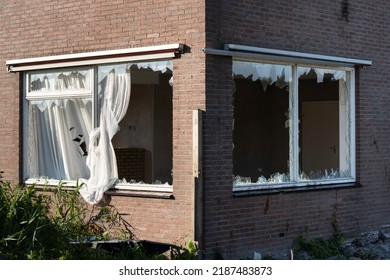 Broken glass windows, caused by vandalism or demolition, in a brick wall of a house, sharp points of glass stick from the rebate. The curtains are partly hanging out