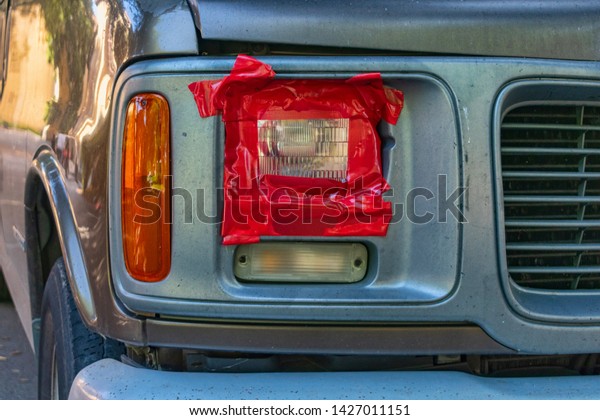 Broken headlight fixed by red duct tape. Car\
accident, insurance concept with adhesive tape on light. Damaged\
headlight detail with temporary tape covering to protect lamp\
against moisture water\
rain.
