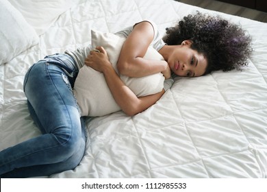 Broken Heart Young Latina Woman Sitting In Bedroom. Depressed Hispanic Girl At Home, Lying On Bed And Holding Pillow With Sad Expression.