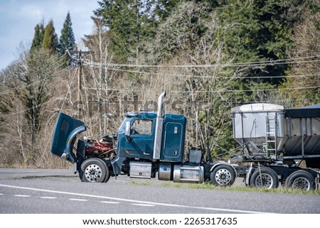 Broken industrial big rig semi truck tractor with an open hood and loaded bulk semi trailer stands on the side of the road waiting for a mobile repair crew or a tow tractor for towing to repair shop