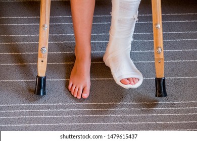 Broken Leg In Cast Of Unrecognizable Little Child.Young Girl In Orthopedic Cast On Crutches In Home. Child With A Broken Leg On Crutches, Ankle Injury. Bone Fracture And Ankle Fracture In Children.