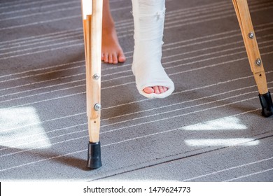 Broken Leg In Cast Of Unrecognizable Little Child.Young Girl In Orthopedic Cast On Crutches In Home. Child With A Broken Leg On Crutches, Ankle Injury. Bone Fracture And Ankle Fracture In Children.