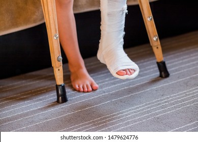 Broken Leg In Cast Of Unrecognizable Little Child.Young Girl In Orthopedic Cast On Crutches In Home. Child With A Broken Leg On Crutches, Ankle Injury. Bone Fracture And Ankle Fracture In Children.