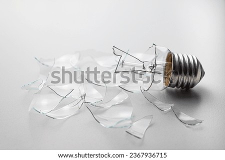 Broken light bulb, with shards of transparent glass around it, on a white surface.