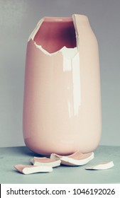 Broken light pink ceramic vase, with fragments in front of it. Grey background