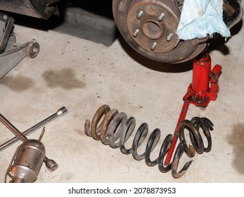 Broken Old Car Coil Spring Parts On Concrete Floor Near A Red Hydraulic Jack And Tools On Wheel Hub Background, Car Suspension Garage Repair