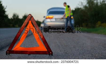 Broken silver luxury car emergency accident. Man driver installing red triangle stop sign on road. Sport automobile turned on blinkers technical problems on the road. Safety procedure vehicle broken