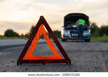 Broken silver luxury car emergency accident. Man driver installing red triangle stop sign on road. Sport automobile turned on blinkers technical problems on the road. Safety procedure vehicle broken