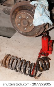 Broken Used Car Coil Spring Parts On Concrete Floor Near A Red Hydraulic Jack And Tools On Wheel Hub Background, Car Suspension Garage Repair