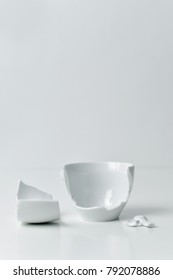 A Broken White Ceramic Coffe Cup With Its Pieces Spread Out On A White Table, Against A White Background With A Blank Space On Top