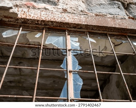 Broken window with rusty metal bars in an abandoned industrial building. Cracked concrete wall, shattered glass and signs of decay in a derelict structure.

