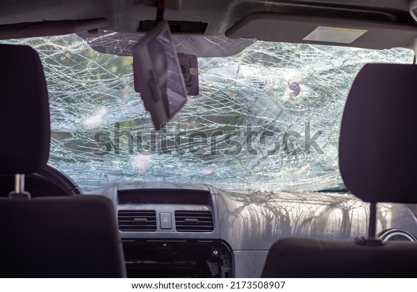 Broken windshield of a car from a bullet, from a\
shot from a firearm, view from the inside of the cabin. Damaged\
glass with traces of an oncoming stone on the road. Ukraine, Irpin\
- May 12, 2022