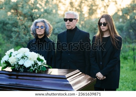Brokenhearted family of three in mourning clothes and sunglasses standing in front of closed coffin with their dead relative or good friend