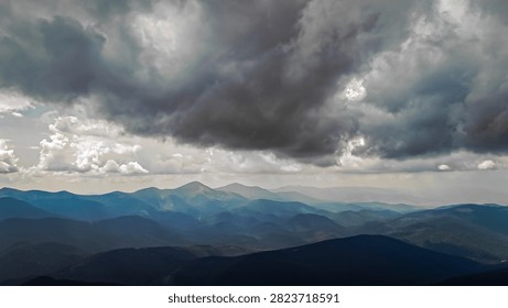Brooding storm clouds loom over layered mountain ranges light filters through the gaps casting silver beams on distant peaks while deep shadows add dramatic mood across the vast wilderness