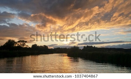 Brooding sunset as darkness falls on the River Nene at Wadenhoe, England, with golden and blue sky, angled cloud formation, black trees silhouetted and relected ripples on the water, more sky