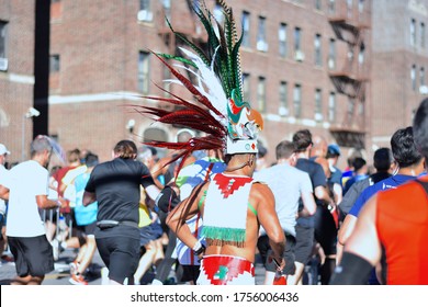 Brooklyn, NY, US - Nov. 4, 2018: A Man In Indigenous Clothing The Color Of The Mexican Flag, Including A Majestic Feathered Headdress, Runs The NYC Marathon In A Crowd Of Runners Seen From Behind.