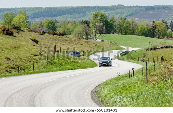 Brosarp, Sweden - May 18, 2017: Documentary of\
Brosarps hills nature area. Car driving the winding road through\
the scenic landscape. Person standing roadside looking through a\
scope at car.