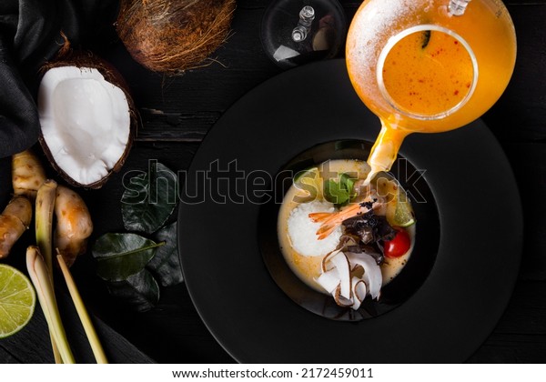 The broth in a jug is poured into a plate with\
rice, shrimps, lime, tomatoes, parsley, tobiko caviar and purple\
lettuce, which is on the table with coconut, rose leaves, black\
cloth, ginger and celery