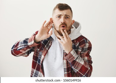 Brother Feeling Insecure And Worried Overhearing Uncomfortable Conversation. Portrait Of Anxious Confused Young Caucasian Man Holding Fingers On Lip And Hand Near Ear, Gossiping And Being Curious