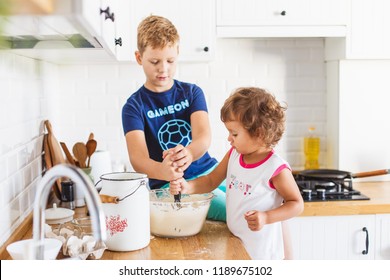 Brother and sister preparing dough for pancakes at the kitchen. Concept of food preparation, white kitchen on background. Casual lifestyle photo series in real life interior