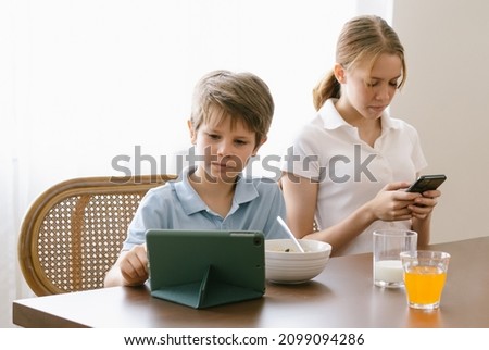 Brother and sister sitting in kitchen during breakfast. Little boy looks at digital tablet and teen girl checking social media holding smartphone. Devices overuse,addiction gadgets dependency concept.