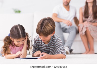 Brother and sister using tablet pc together on floor with parents behind them - Powered by Shutterstock