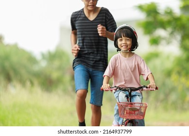 Brother Teaching Sister Practice Cycling Looked After Him With Concern. Riding An Outdoor Exercise Bike Is An Outdoor Activity That Enhances Family Holiday Learning. Background Is Green Trees.