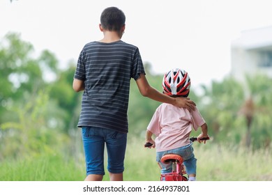 Brother Teaching Sister Practice Cycling He Looked After Him With Concern. Riding An Outdoor Exercise Bike Is An Outdoor Activity That Enhances Family Holiday Learning. Background Is Green Trees.