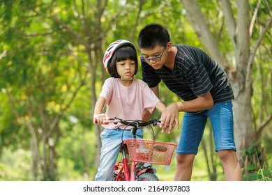 Brother Teaching Sister Practice Cycling Looked After Him With Concern. Riding An Outdoor Exercise Bike Is An Outdoor Activity That Enhances Family Holiday Learning. Background Is Green Trees.