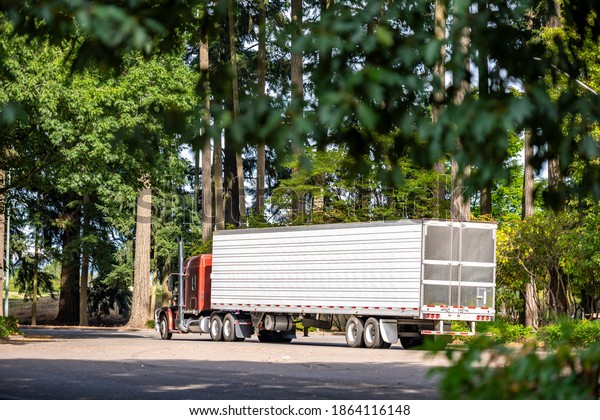 Brown big\
rig industrial semi truck tractor with loaded refrigerator semi\
trailer carry frozen food for delivery and running on the rest area\
parking lot with old trees to highway\
entrance