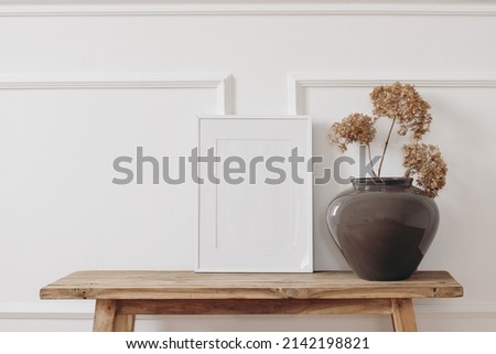 Brown bold vase. Dry hydrangea flowers on old wooden bench. Blank black picture frame mockup. White wall moulding background, stucco decor. Empty copy space. Elegant interior. Summer, fall still life 