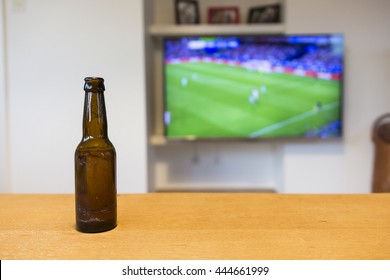 A Brown Bottle Of Beer Standing On A Wooden Table. A Soccer Match On Tv Is Visible In The Background, Which Is Blurred Out.