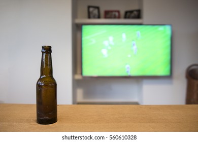 A Brown Bottle Of Beer Standing On A Wooden Table. A Soccer Match On Tv Is Visible In The Background, Which Is Blurred Out.