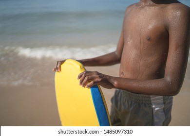 Brown boy holding yellow surf board with beach waves on the background.