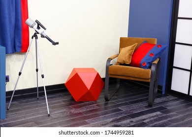 A Brown Chair With Two Pillows. Bright Tones Of The Children's Room, Red, White, Blue, Brown. Children's Toy, Built-in Duffcase Cabinet. Telescope For The Study Of Stars.