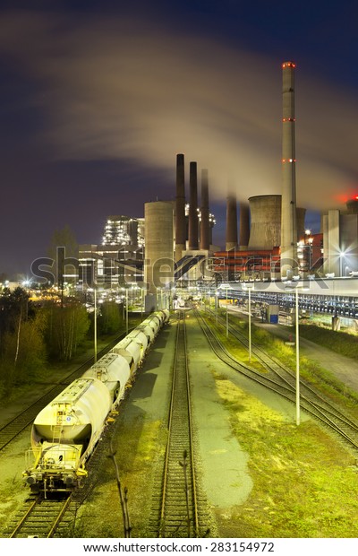 A brown coal power station with night\
blue evening sky and a train in the\
foreground.
